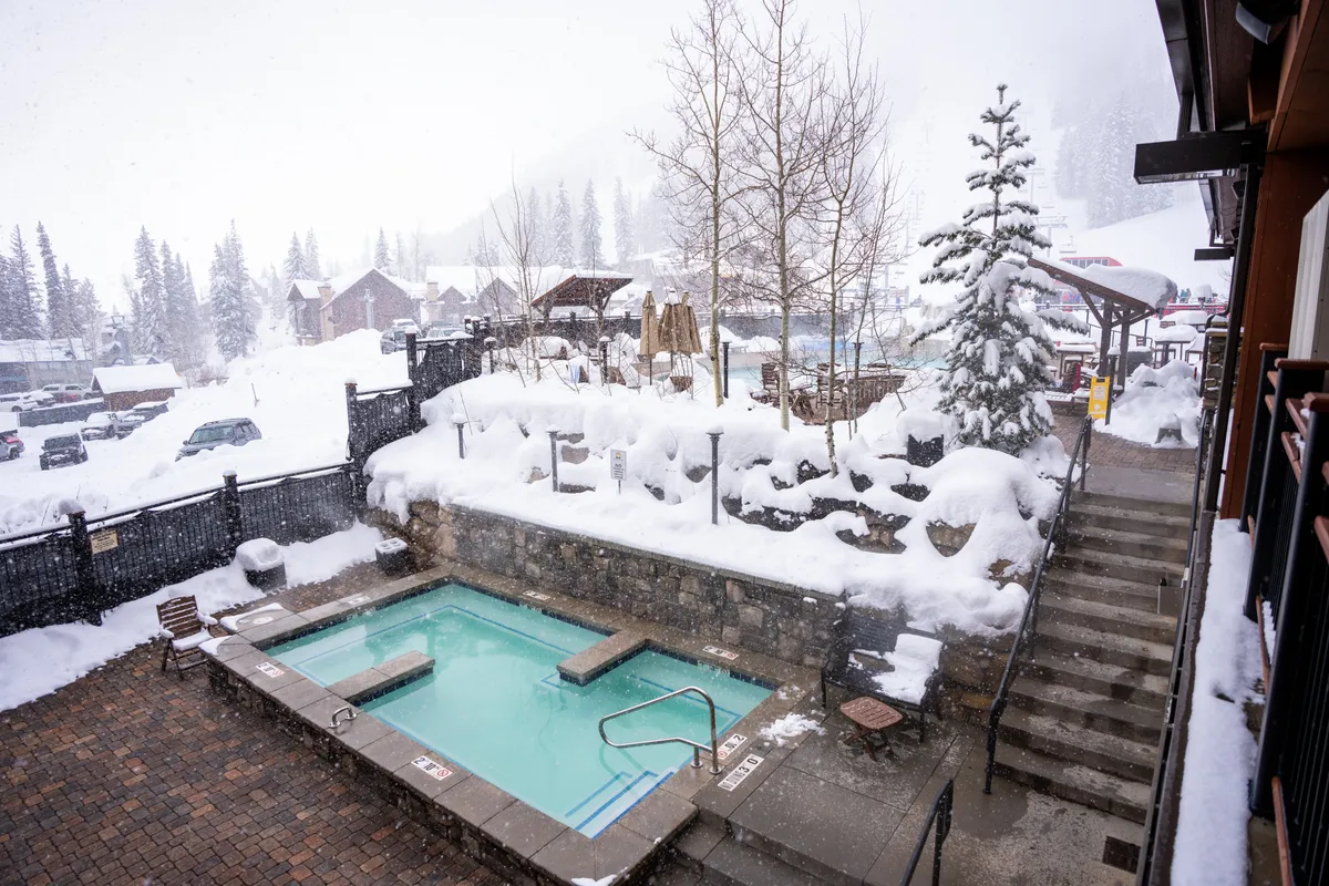 This exterior image features a steaming hot tub set on a terrace, surrounded by snow-covered trees. Guests are soaking and socializing, enjoying the contrast of warmth and winter. The snow falls gently, creating a tranquil, alpine atmosphere that epitomizes après-ski relaxation.