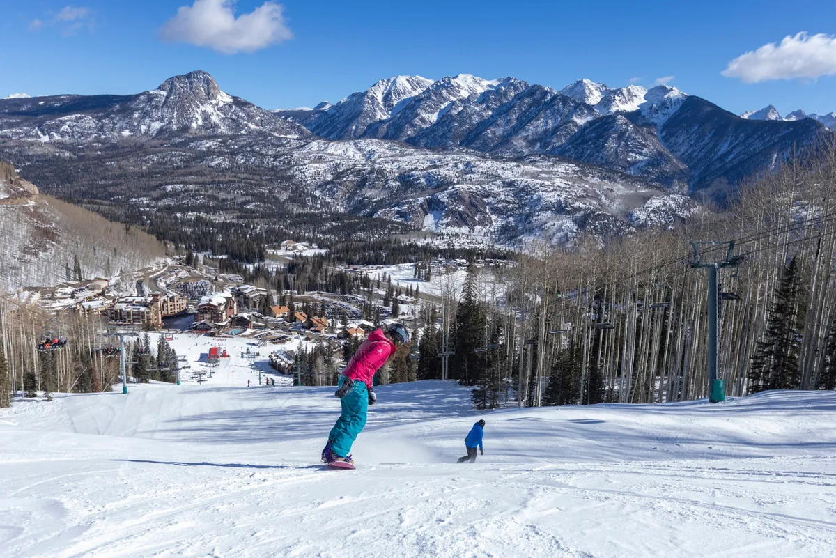 This vibrant exterior shot captures a skier and child enjoying the pristine, snow-covered slopes with a majestic mountain range and ski resort buildings in the background. The blue sky and fresh snow evoke a sense of adventure and family fun at the heart of the ski-in, ski-out experience.