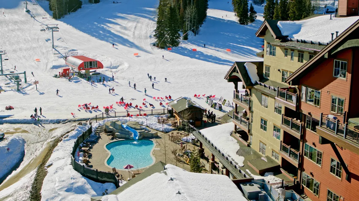 An inviting outdoor swimming pool sits at the base of the ski slopes, with people relaxing by the pool and others skiing in the background. The lodge and ski lifts are visible, highlighting the seamless blend of resort amenities and mountain activities.