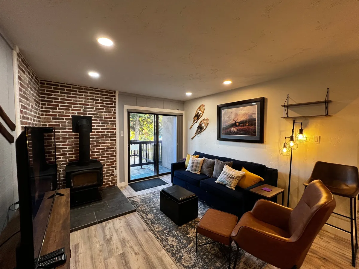 A stylish and cozy living area with a brick accent wall, wood-burning stove, plush sofa adorned with decorative pillows, and a sliding glass door leading to a balcony. Soft natural light fills the room, highlighting a welcoming space for gatherings.