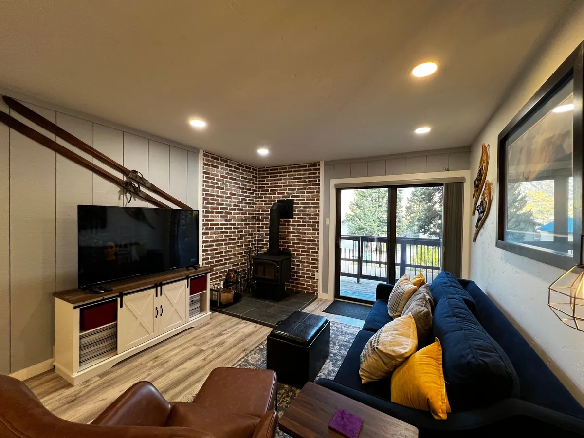 A warm and inviting living room, featuring a sofa with colorful cushions, a brick wall with ski-themed decor, a modern television unit, and a cozy wood stove. A large window reveals a scenic mountain view, bathing the space in natural light.