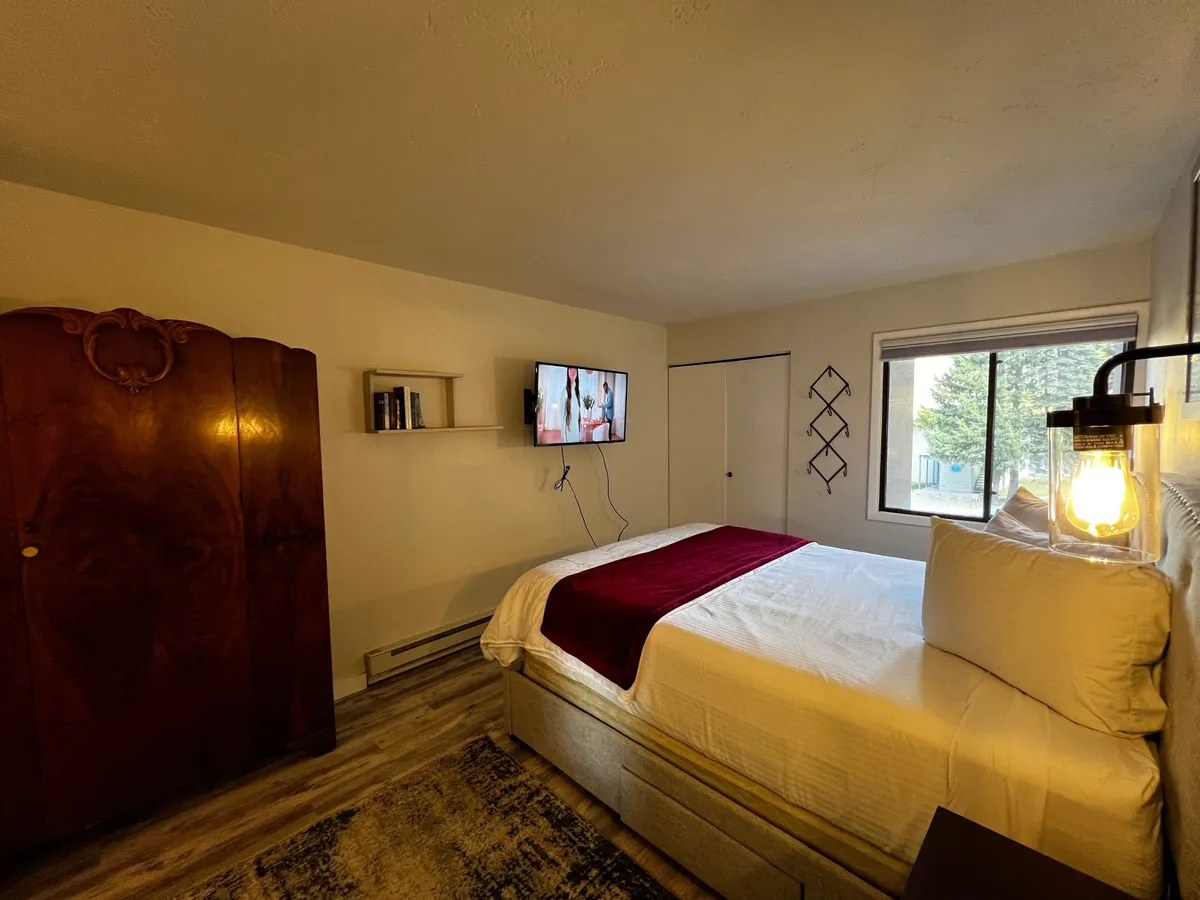 A cozy queen bedroom featuring a bed with a burgundy throw, wall-mounted TV, and a wooden wardrobe. Natural light filters in, enhancing the inviting atmosphere and highlighting the restful space ideal for relaxation after a day outdoors.