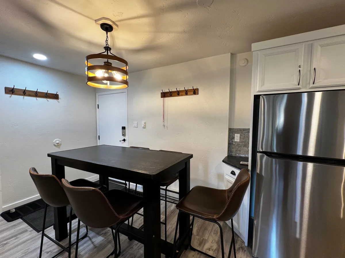 A cozy dining area anchored by a sturdy dining table beneath a soft-glowing chandelier. The refrigerator in the background and comfortable seating for a group highlight the space’s capacity for family-style dining and lingering over drinks or games.