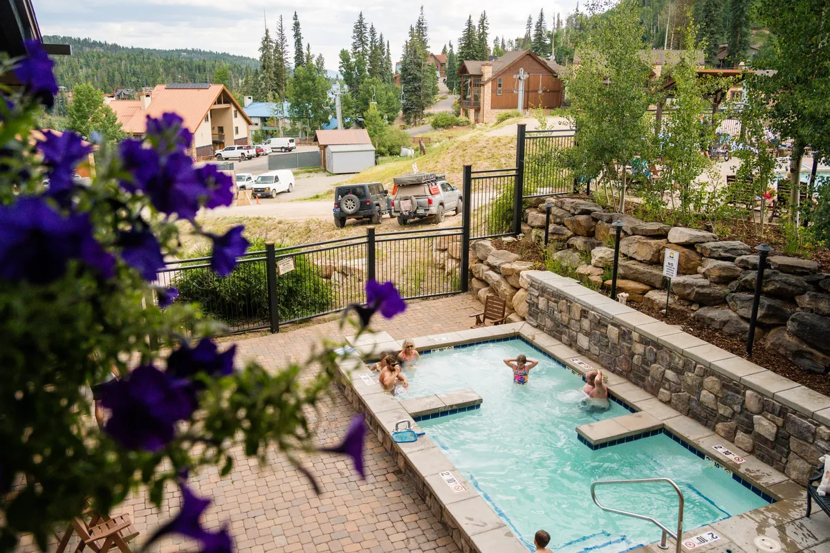An inviting outdoor hot tub with people relaxing, surrounded by lush greenery and a beautiful mountain view in the background. The setting is tranquil, with a stone wall edging the patio area and hints of sunset light.