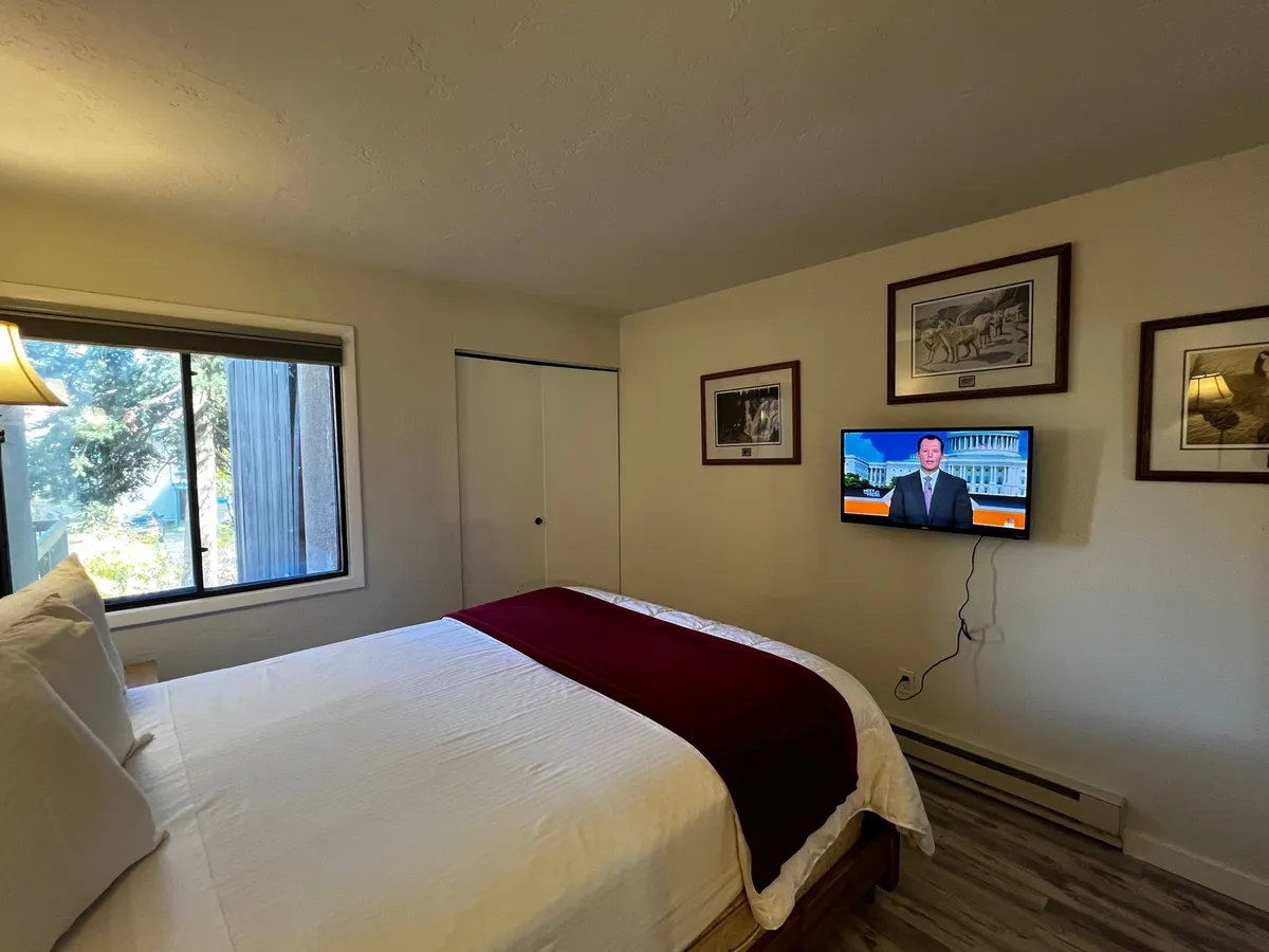 A queen bedroom with white linens accented by a maroon blanket, a large window that lets in natural light, a mounted television, and artistic wall décor. The ambiance feels inviting and peaceful, echoing the restful retreat described.