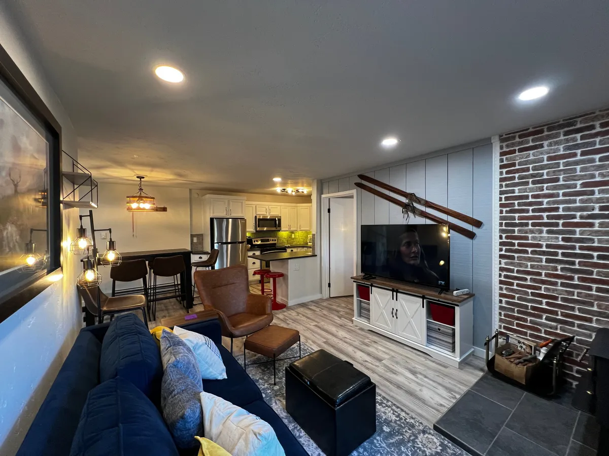 A spacious interior with a blue sleeper sofa, wooden dining table with chairs, a television unit adorned with decorative objects, and an exposed brick wall. The kitchen area is visible, creating a sense of open-plan living ideal for group activities.