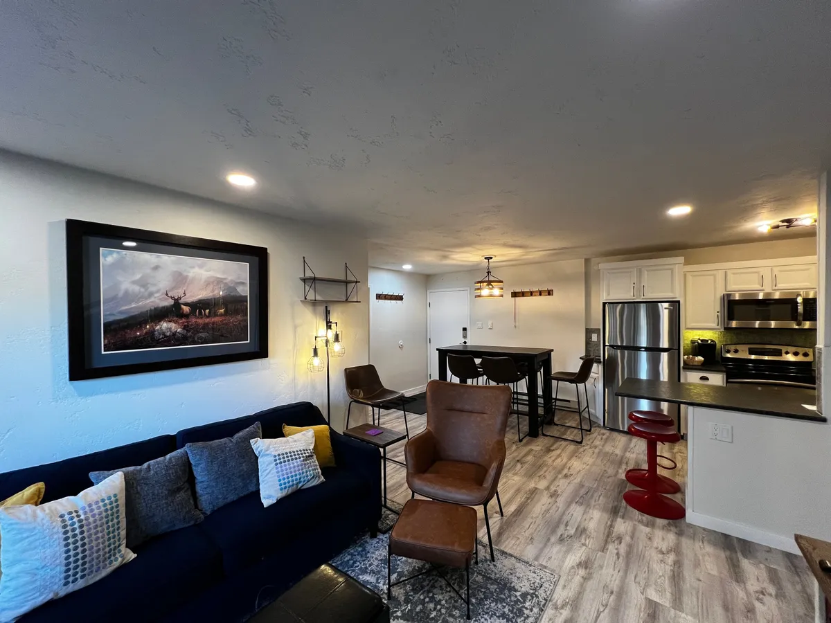 The hero image shows a welcoming open-concept living space featuring a cozy sofa with colorful pillows, stylish framed artwork on the walls, a modern dining table perfect for family meals, and a well-equipped kitchen area. The overall atmosphere is warm, inviting, and ideal for relaxing or spending time together after a day on the slopes.